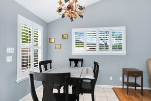 A dining area with a table and chairs, two windows with blinds, a chandelier, and wall-mounted light switches.