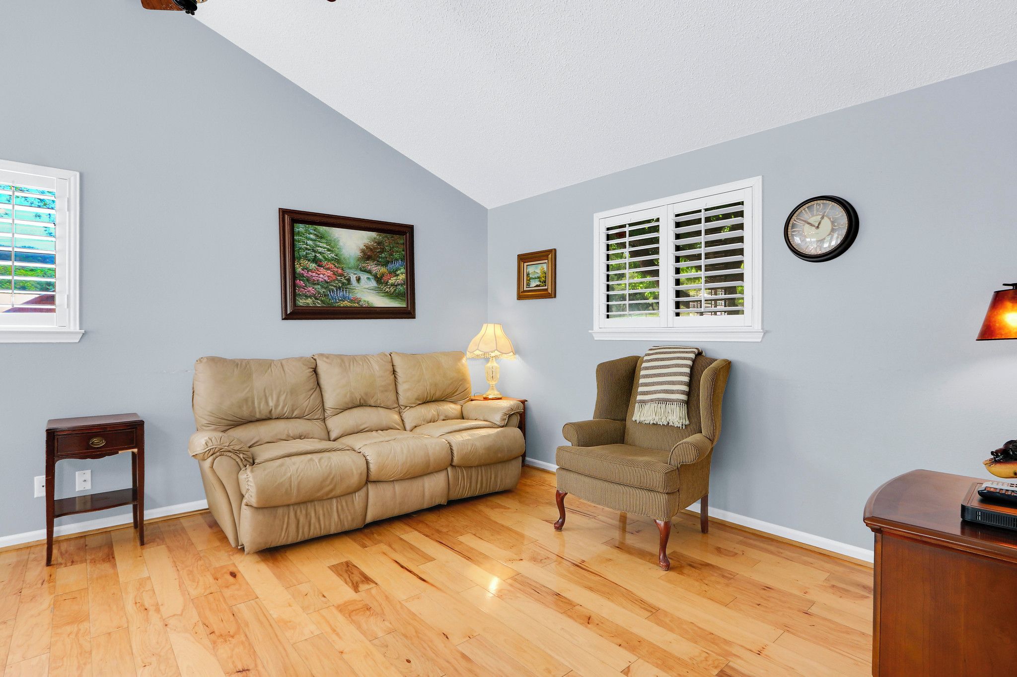 A living room with a beige sofa, a striped armchair, a small table, a lamp, and a clock on the wall.
