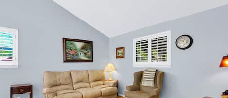 A living room with a beige sofa, a striped armchair, a small table, a lamp, and a clock on the wall.