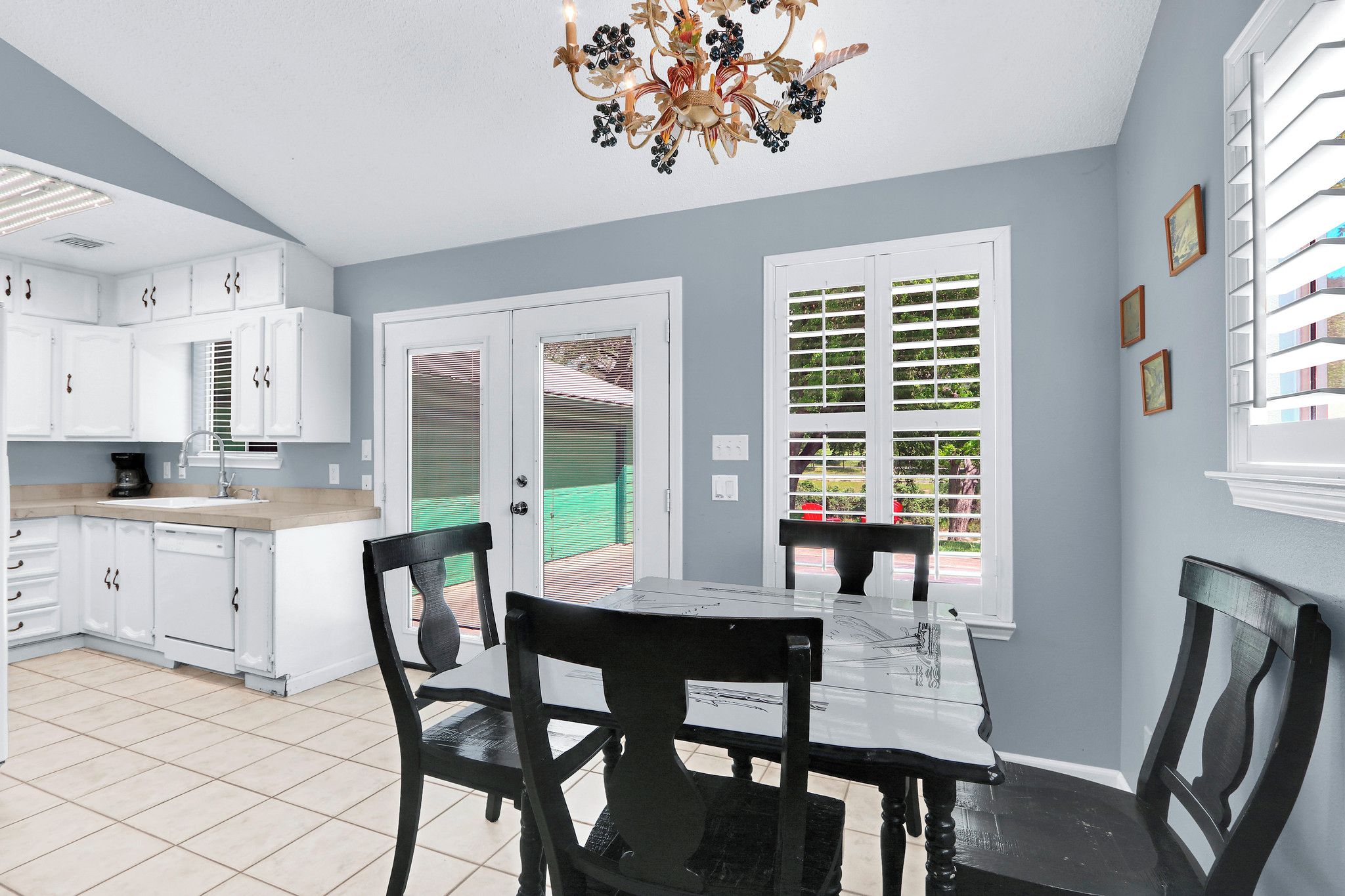 A kitchen with white cabinets, a dining table with chairs, a chandelier, and a view through a window with blinds.