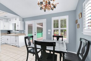 A kitchen with white cabinets, a dining table with chairs, a chandelier, and a view through a window with blinds.