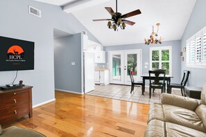 A spacious living room with a wooden floor, a ceiling fan, a television, and a dining area with a table and chairs.