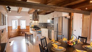 Una cocina moderna con vigas de madera, armarios blancos y un comedor listo para seis personas, con fruteros y servilletas.