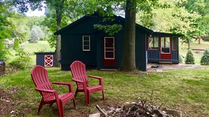 A cabin with two red Adirondack chairs in front, surrounded by trees and a grassy yard.