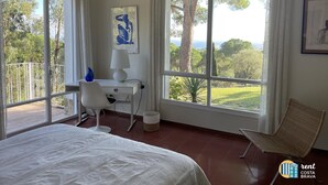 Un dormitorio con cama, escritorio, silla y ventanas amplias que dan a vistas de árboles y al mar.