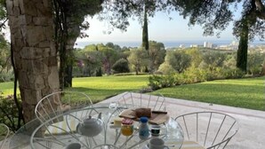 Una mesa redonda de cristal con sillas, una tetera, tazas y una botella, ubicada al aire libre con vistas a un paisaje verde y edificios a lo lejos.