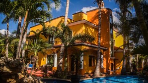 Un edificio amarillo con piscina, rodeado de palmeras.