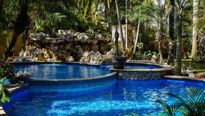 Una piscina con cascada de roca, rodeada de plantas tropicales y un muro de piedra.