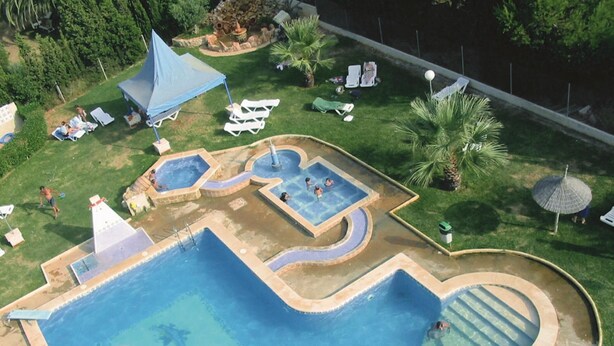 A large outdoor swimming pool with multiple sections, surrounded by lounge chairs and palm trees. - APCOSTAS San Antonio / One Bedroom (Alcoceber)