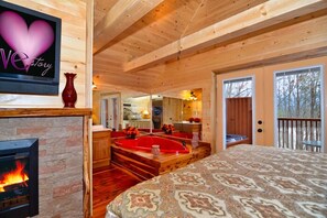 A cozy cabin interior with a fireplace, a large bed, a red jacuzzi, and a TV displaying 'Love Story'.