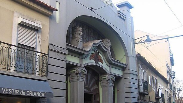 A building with a large archway entrance, a sign for 'VESTIR de CRIANÇA', and a person sweeping the sidewalk.