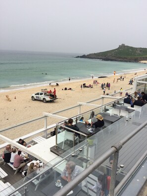 A beachside restaurant with outdoor seating overlooking the ocean, a lifeguard vehicle, and a clear view of the beach and sea. - St. Ives Garden apartment (CORNWALL)