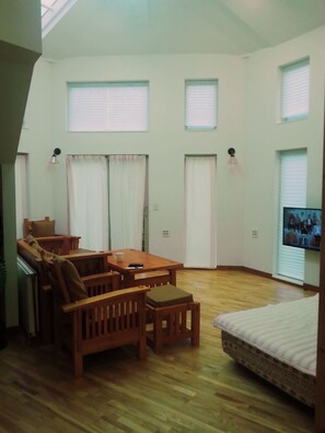 Una habitación amplia con una mesa de comedor de madera, sillas, un sofá y una cama. Hay tres ventanas con persianas, un ventilador de techo y un televisor montado en la pared.