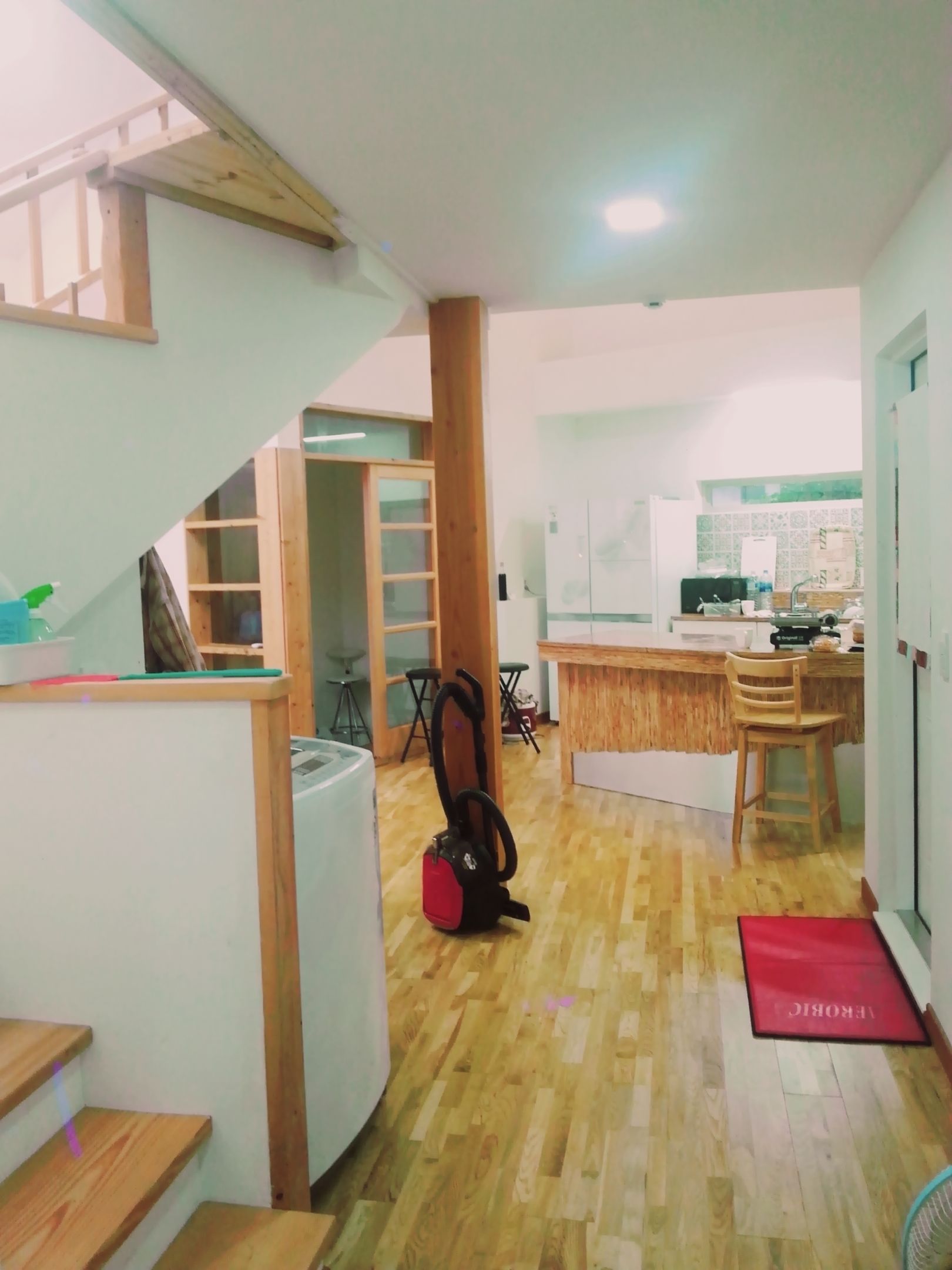 A kitchen with wooden flooring, a red mat at the entrance, and a staircase leading to another room.