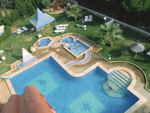 Una amplia piscina al aire libre con varias secciones, rodeada de sillas de descanso y palmeras.