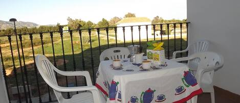 A balcony with a table set for two, featuring a tablecloth with a tea set design, white plastic chairs, and a view of a grassy landscape with trees and hills in the background.