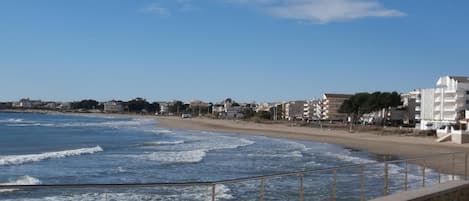 A coastal promenade with a stone railing, beachfront buildings, and a clear blue sky.