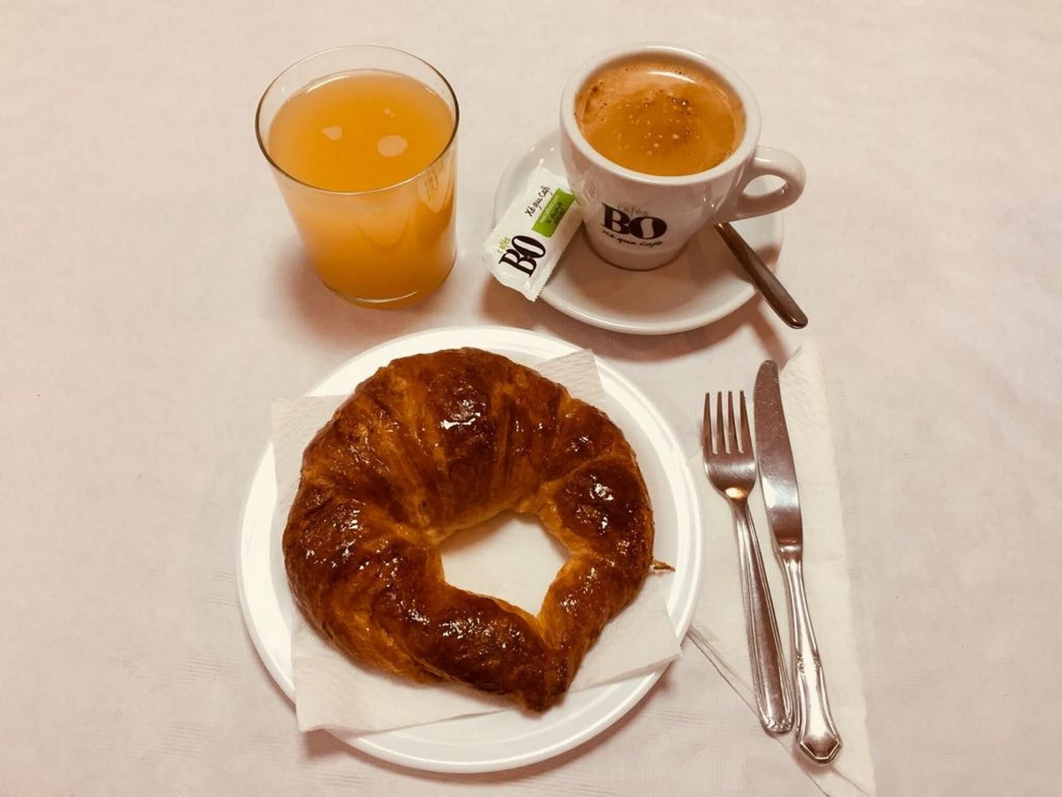 En frukostuppdukning med en croissant, en kopp kaffe och ett glas apelsinjuice på ett vitt bord.