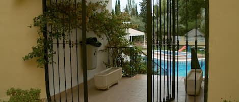 A gated outdoor area with a pool, potted plants, and a white building in the background.