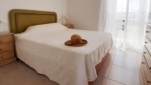 Un dormitori amb llit, una tauleta de nit, una làmpada, una petarrera i una finestra amb cortines translúcides.