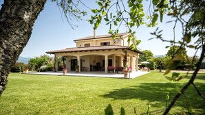 Ein zweistöckiges Haus mit überdachter Terrasse, umgeben von einem gepflegten Rasen und Kübelpflanzen.