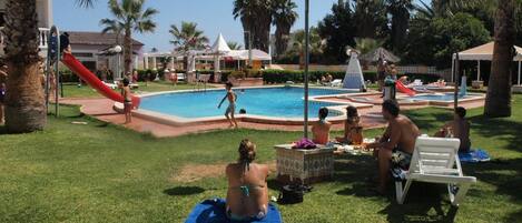 A pool area with a slide, palm trees, and people relaxing on towels.