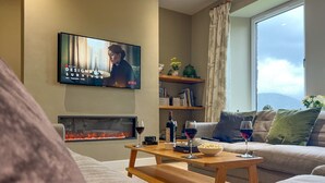 A living room with a flat-screen TV, a fireplace, a coffee table with snacks and drinks, and a large window with a view. - Lake View, luxurious home with 2 ground floor bedrooms, stunning Lake Ullswater views. (Penrith)