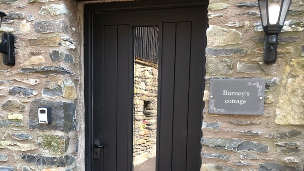 Un bâtiment en pierre avec une porte noire, une plaque portant l’inscription « Barney’s cottage », et un luminaire fixé au mur.