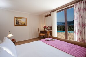 A hotel room with a bed, a desk, a chair, a lamp, a painting, and a view of the sea through large windows. - Luxury villa overlooking the sea with private pool-Villa Rose (Lefkada)