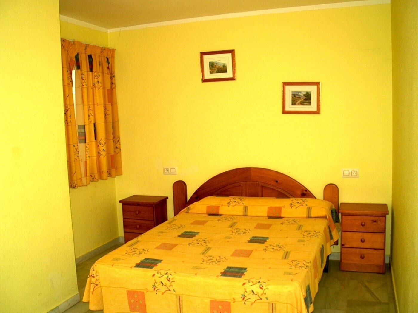 Une chambre à coucher avec un lit en bois, deux tables de chevet, une fenêtre avec des rideaux et deux tableaux encadrés accrochés au mur.