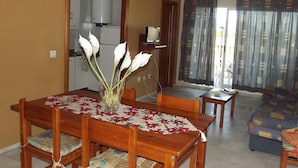 A dining area with a wooden table, chairs, and a vase of flowers. There is a kitchen in the background and a living area with a sofa and curtains. - APCOSTAS San Antonio / Three Dedrooms (Alcoceber)