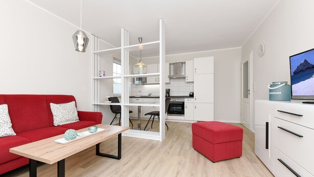 A modern living room with a red sofa, a wooden coffee table, and a red ottoman. A kitchen with white cabinets and a dining area with a table and chairs is visible in the background. - Sandpiper - Haus Andreesen (Neuharlingersiel)