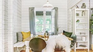 Un comedor acogedor con una mesa de madera, sillas y una butaca blanca con un plaid de piel sintética. Se ve una estantería blanca con cristalería y una planta.