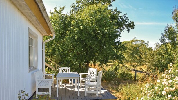 Un patio in legno con tavolo e sedie, immerso nel verde e affiancato da un edificio bianco.