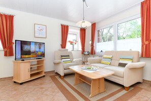 Ein Wohnzimmer mit Fernseher, Sofa, Sessel, Couchtisch und Fenstern mit Vorhängen.
