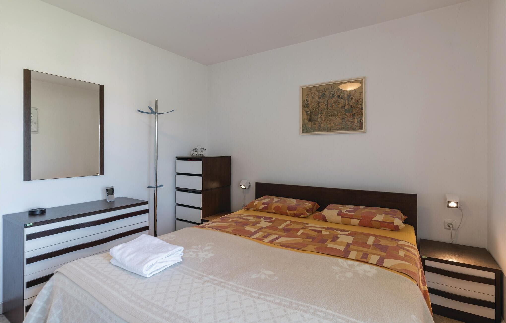 Een slaapkamer met een bed, een dressoir, een spiegel, een kapstok, een nachtkastje en een wandlamp.