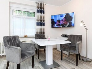 Un salon moderne avec une table blanche, des fauteuils gris, un téléviseur à écran plat et un lampadaire.