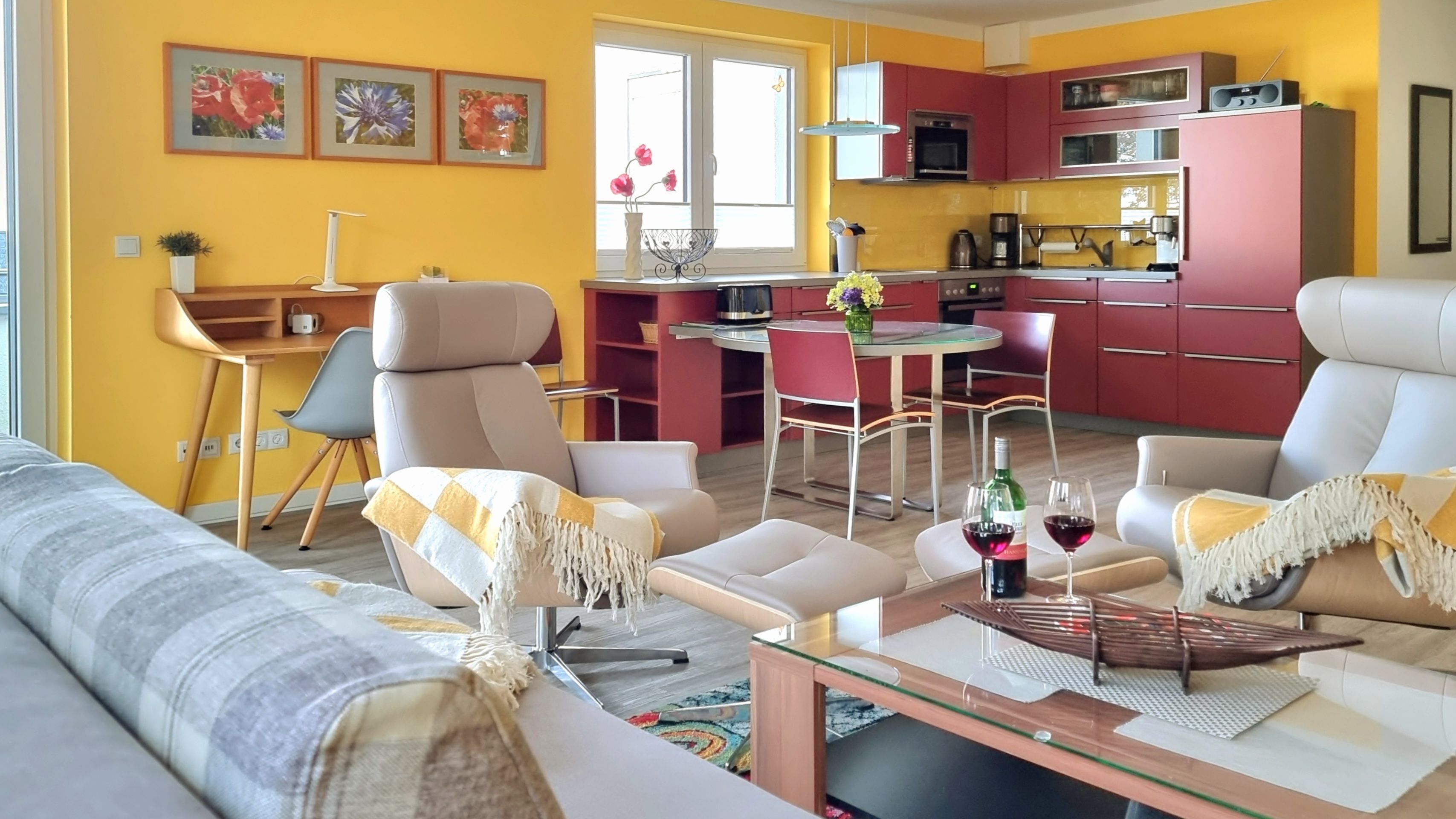A modern living room with a sofa, armchairs, and a coffee table. There is a dining area with a table and chairs. The kitchen is visible with red cabinets and a microwave.