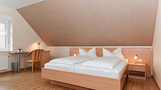 Een slaapkamer met schuine balken, een houten bed, een nachtkastje met lamp, een stoel en een bureau.