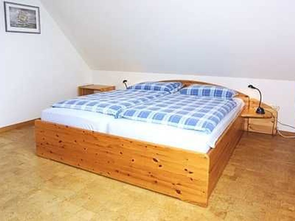 Een enkel bed met houten frame en een blauw-witte geruite spreidekker in een kamer met witte muren en een schuine plafond.