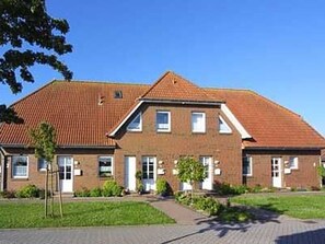 Ein zweistöckiges Backsteingebäude mit Ziegeldach, einem zentralen Eingang und einer kleinen Grünfläche davor.