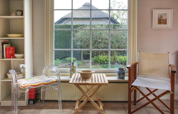 Een kamer met een groot raam, een houten tafel, een transparante stoel, een houten stoel en een boekenkast met boeken en decoratieve objecten.