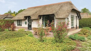 Une maison traditionnelle à toit de chaume, avec une façade en briques, une porte d’entrée et un petit jardin agrémenté d’une table et de chaises.