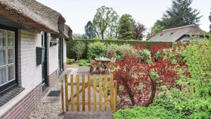 Un bâtiment à toit de chaume, doté d’un portail en bois, un jardin aux feuillages rouges et une table avec des chaises à l’extérieur.