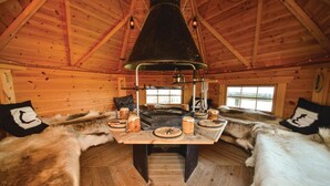 L’intérieur d’une cabane en rondins, avec un foyer central, des bancs recouverts de fourrure et des murs en bois.