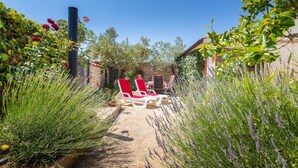 Un sendero en un jardín conduce a una zona de estar con sillas rojas y una mesa, rodeada de abundante vegetación y plantas de lavanda.