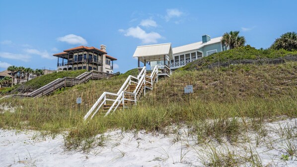 A beachfront property with a staircase leading to a white building with a red roof, surrounded by sand and vegetation.