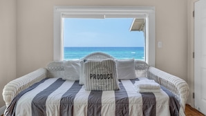 Een slaapkamer met een groot raam met uitzicht op de oceaan, een bed met gestreept beddengoed en een kussen met de tekst 'MARKET FRESH LOCAL PRODUCE ORGANIC SINCE 1871'.