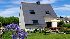 Una casa moderna con techo inclinado, un pequeño balcón con mesa y sillas, y un jardín con flores moradas.