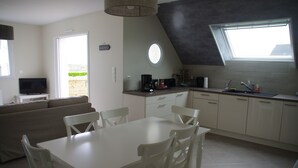 Una cocina moderna con armarios blancos, una mesa de comedor y sillas. Hay un televisor en la sala de estar y una ventana que proporciona luz natural.